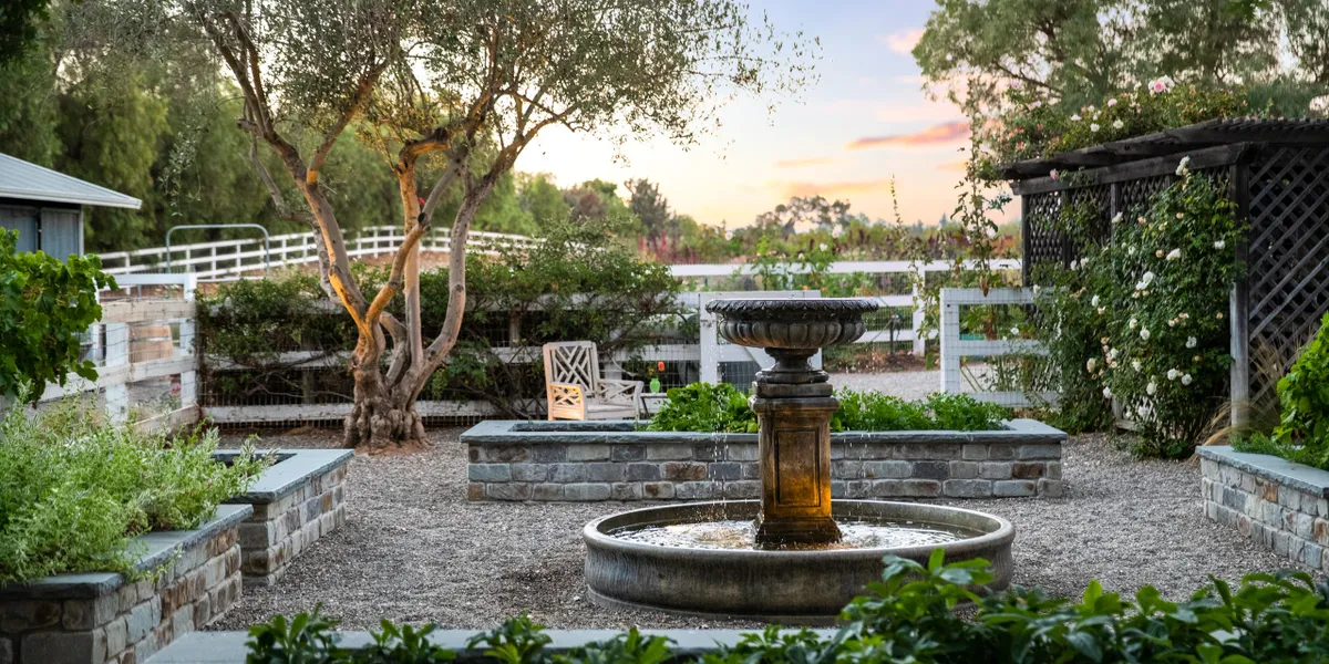 Fountain | AAA Landscaping Elegant backyard landscaping with a rustic stone fountain, lush greenery, and a peaceful garden setting at sunset, creating a serene outdoor retreat.